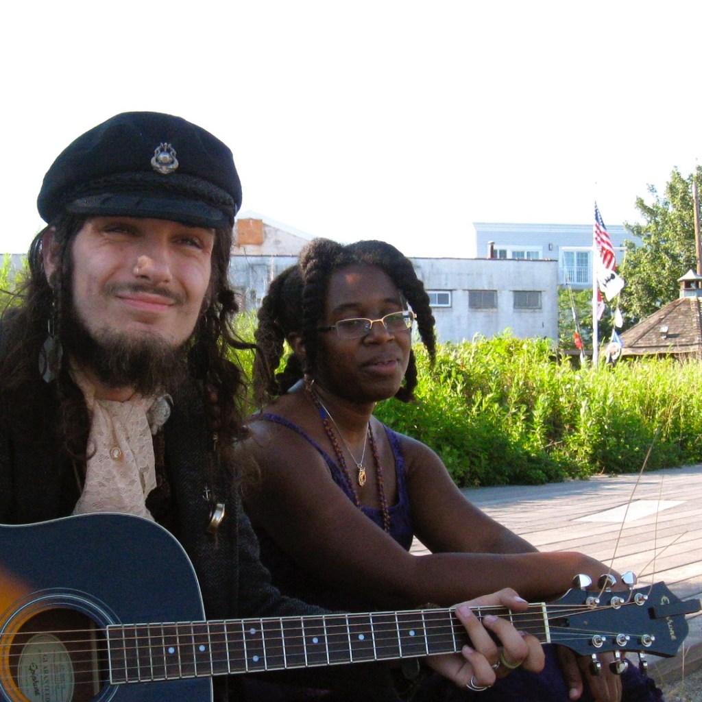 Sam playing guitar and Krystal singing while sitting on a boardwalk